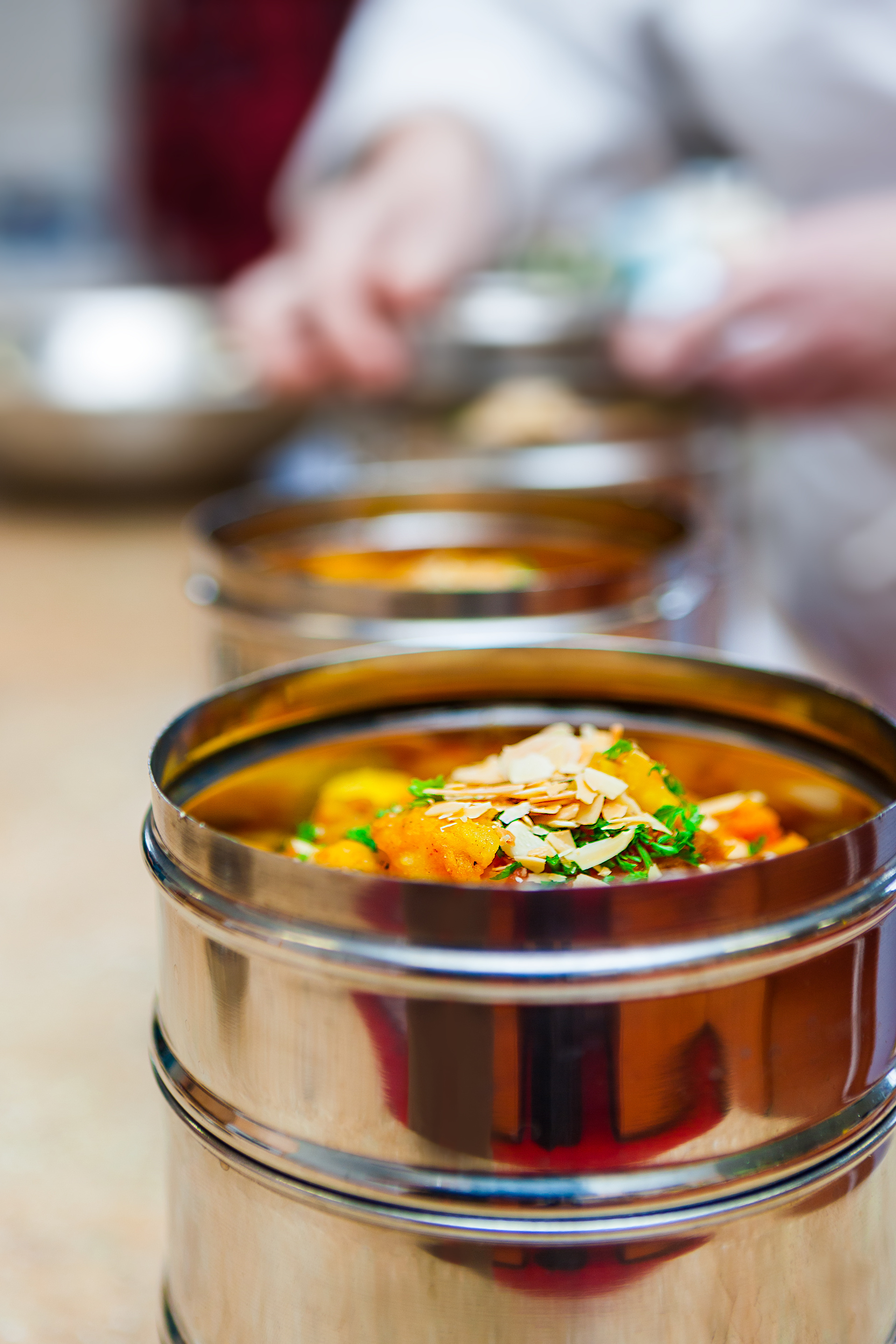 Macro Close Up of the Top of Stainless Steel Tiffin (Dabba) Container with Curried Vegetable Stew and Chef's Hands in the Background